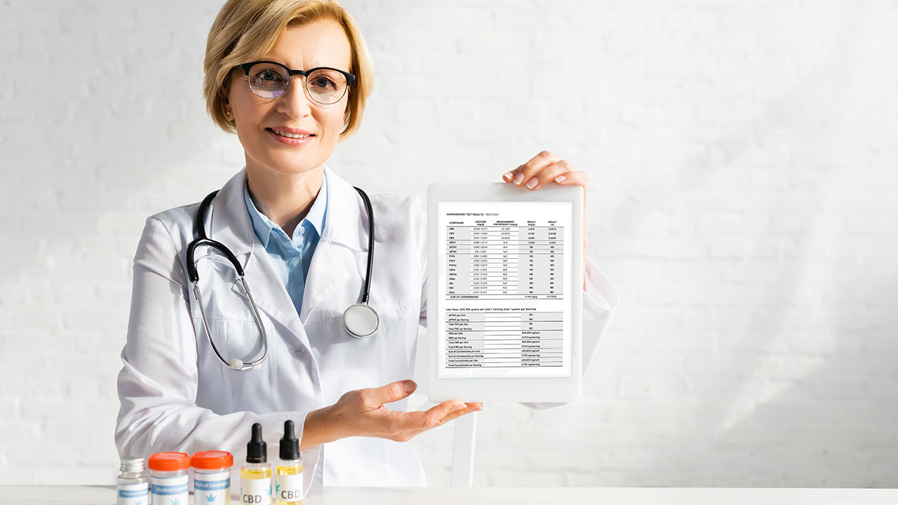 A woman with short blonde hair in a lab coat with a stethoscope  holding up a COA report on a tablet with bottles of CBD products in front of her.