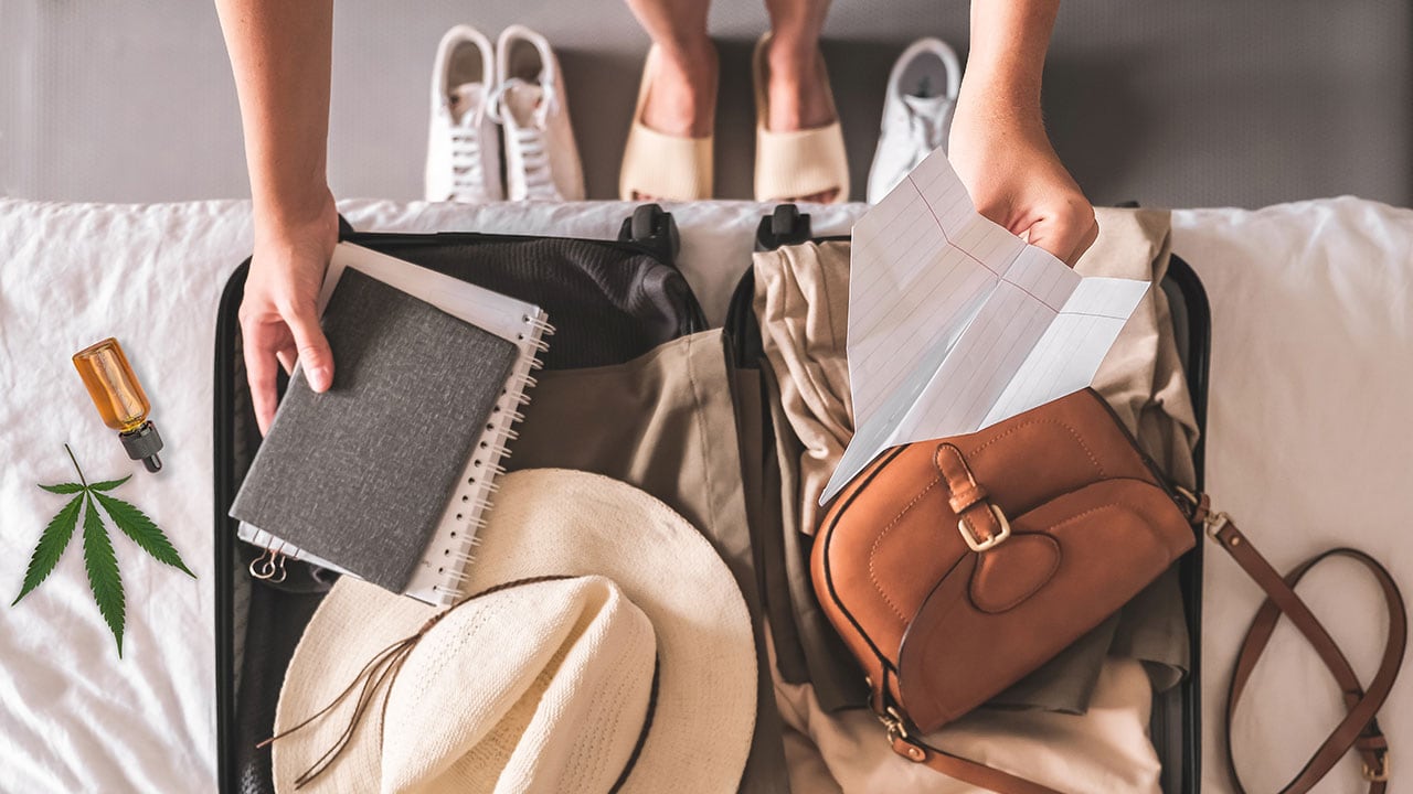 A person reaching over an open suitcase on a bed holding a paper airplane and notebook with a bottle of CBD oil next to the suitcase