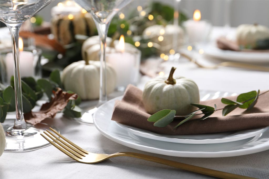 Close-up of a festive Thanksgiving table featuring a white pumpkin, greenery, candles, and gold flatware, creating a warm, welcoming atmosphere for celebrating Danksgiving traditions.