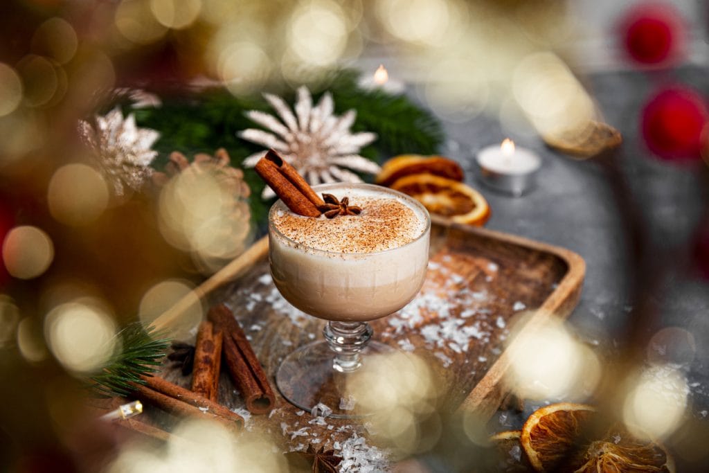 Creamy holiday eggnog served in a glass with cinnamon sticks and star anise, surrounded by dried oranges, pine greenery, and soft candlelight — a warm, seasonal setting ideal for a cannabis-infused twist.