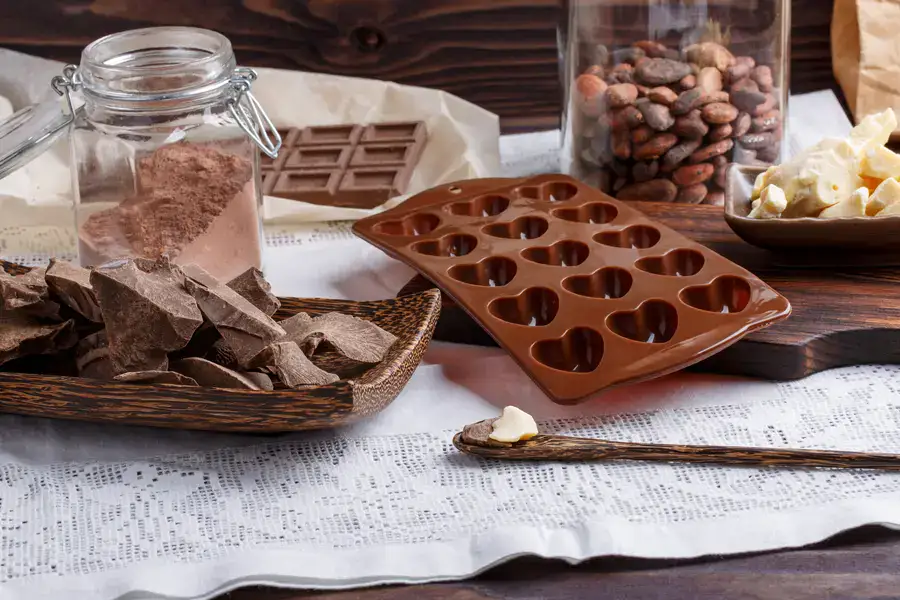 Chocolate-making ingredients and tools including dark chocolate pieces, cocoa powder, cocoa beans, butter, and silicone heart molds on a wooden surface