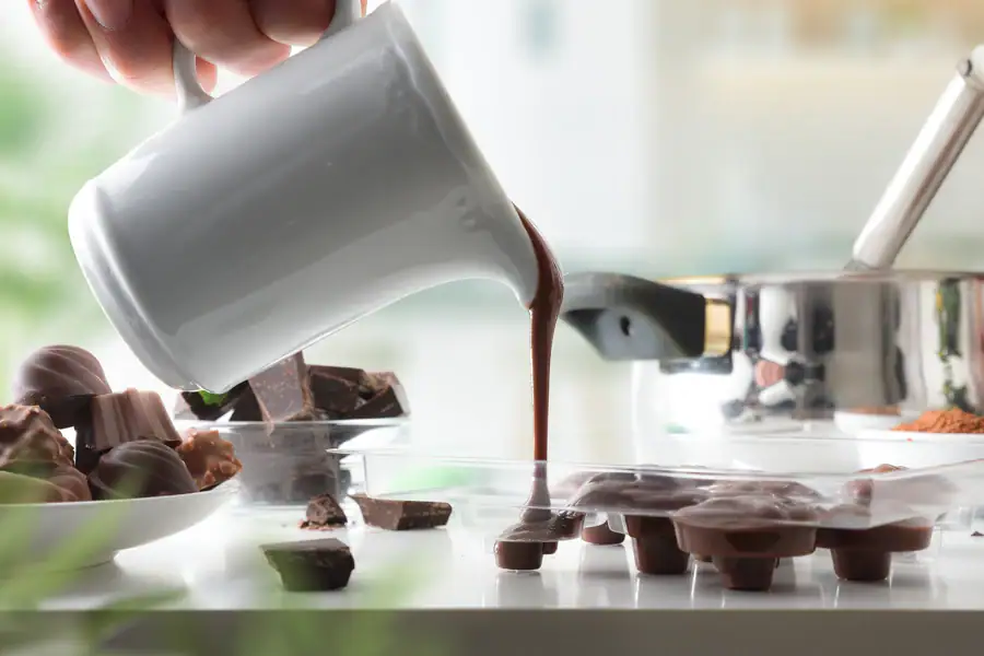 Melted chocolate being poured from a small pitcher into silicone molds on a kitchen counter