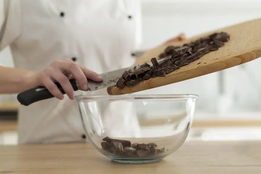 Chopped dark chocolate being transferred from a cutting board into a glass bowl for melting