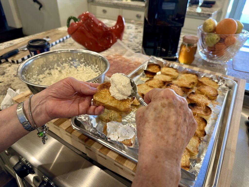 Hands spreading fresh ricotta onto toasted crostini in a kitchen, with a bowl of ricotta mixture and a tray of toasted bread slices in the background.