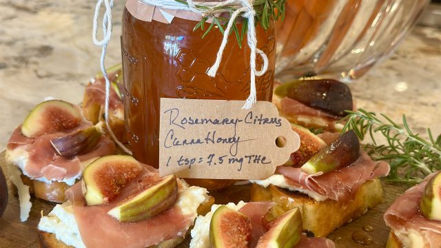 Jar of homemade Rosemary Citrus CannaHoney labeled with dosing information, surrounded by toasted crostini topped with ricotta, prosciutto, fresh figs, and rosemary on a wooden board.